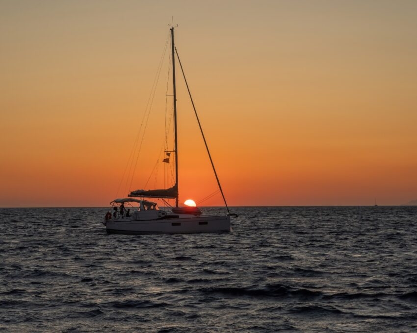 A sailboat on the ocean against a sunset with the sun partially obscured by the boat.