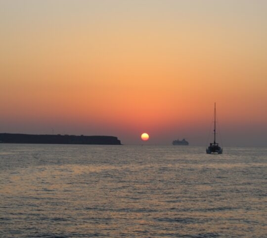 Sunset over the sea with silhouette of a sailboat and distant land.