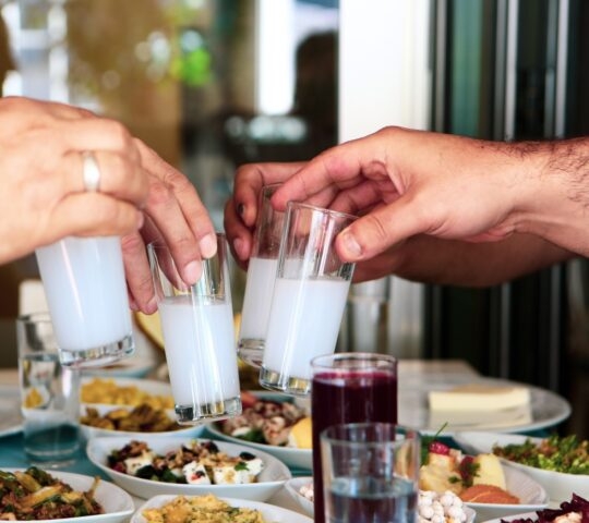 Two hands toasting with glasses of white beverage over a table filled with various dishes.