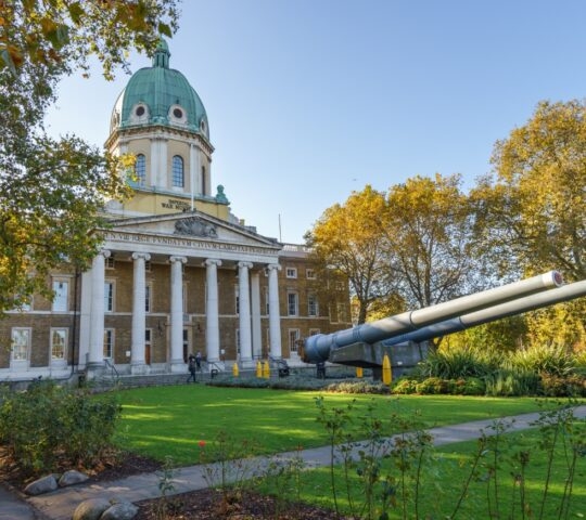 Classical building with a dome and a large cannon in front, surrounded by trees and gardens.