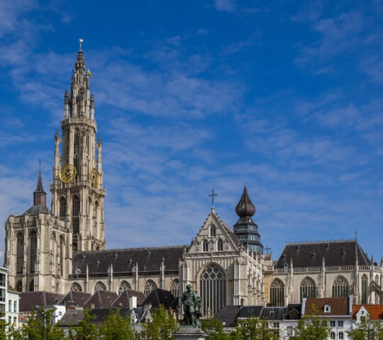 Ornate gothic cathedral with a tall spire, clock faces, against a blue sky.