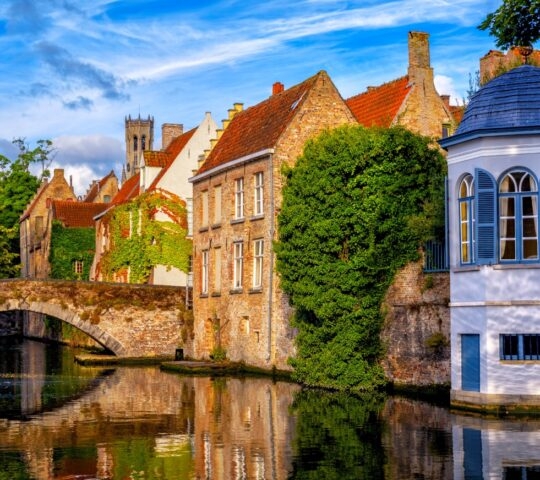 Historic European buildings by a canal with a stone bridge and lush greenery.