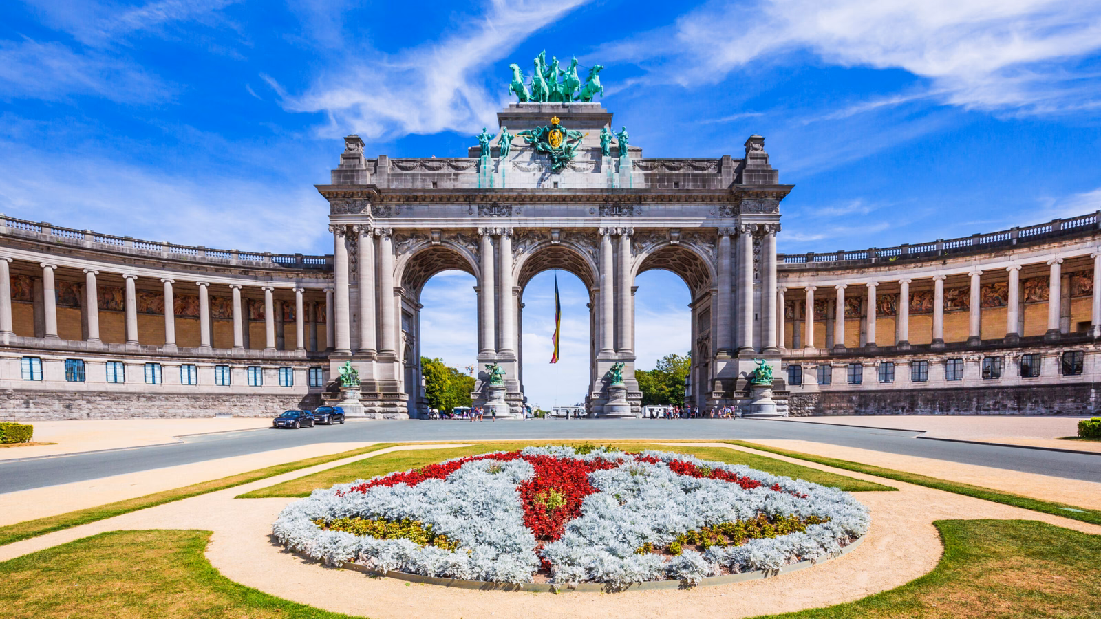 A grand triumphal arch with sculptural details and a colourful floral foreground under a blue sky.