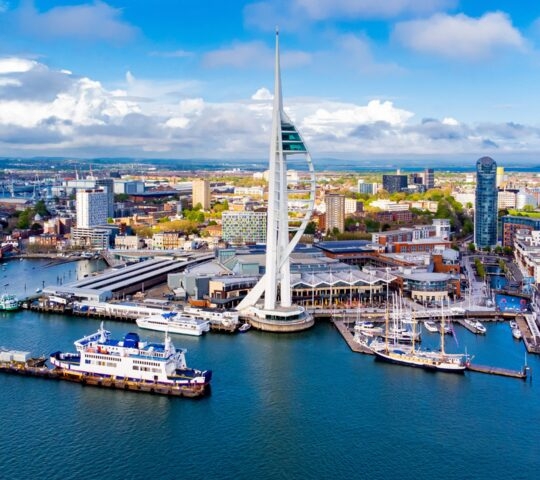 Aerial view of a coastal city with a prominent spire-like tower near the harbor.