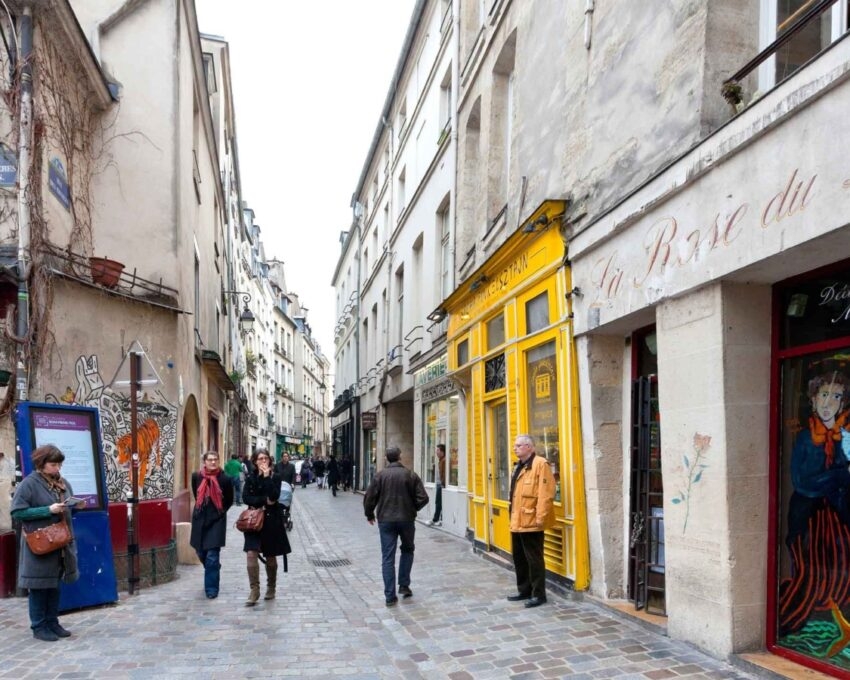 A quaint narrow street with people, vibrant storefronts, and cobblestone paving.