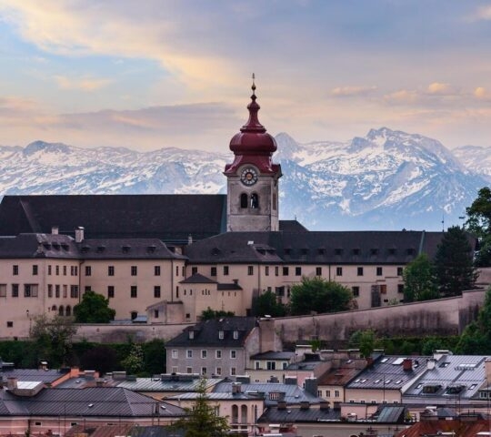 A church with a red spire overlooks a town with snow-capped mountains in the background.