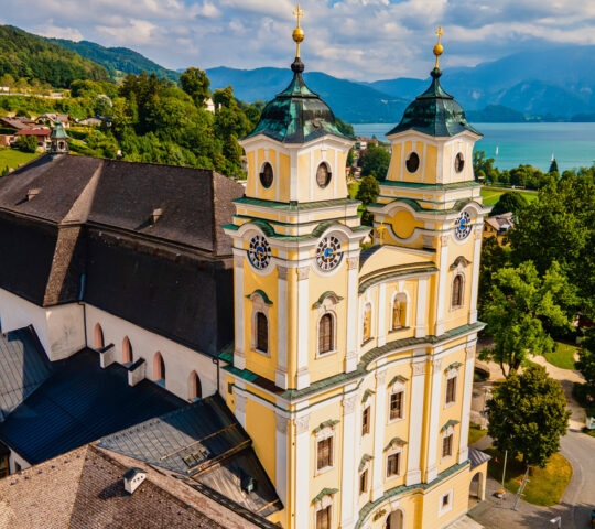 Aerial view of a Baroque church with twin towers overlooking a lake and mountains.