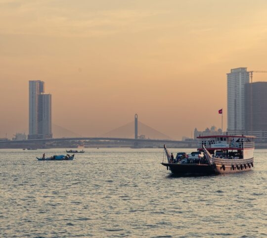 Sunset view of a coastal cityscape with boats on water and a bridge in the distance.