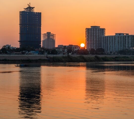 Sunset over a river with silhouette of city skyline and a small boat on the water.