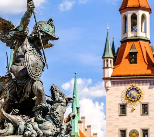 Bronze statue of a winged figure in front of a clock tower with a blue sky background.
