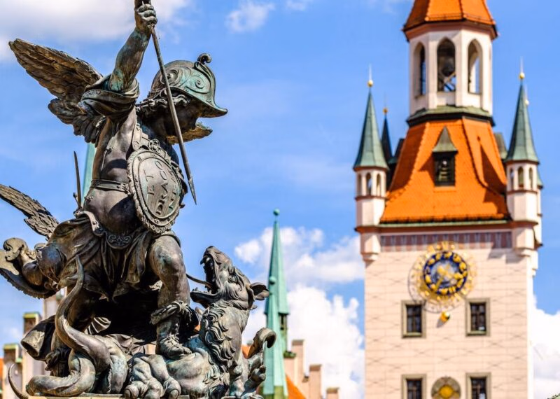 Bronze statue of a winged figure in front of a clock tower with a blue sky background.