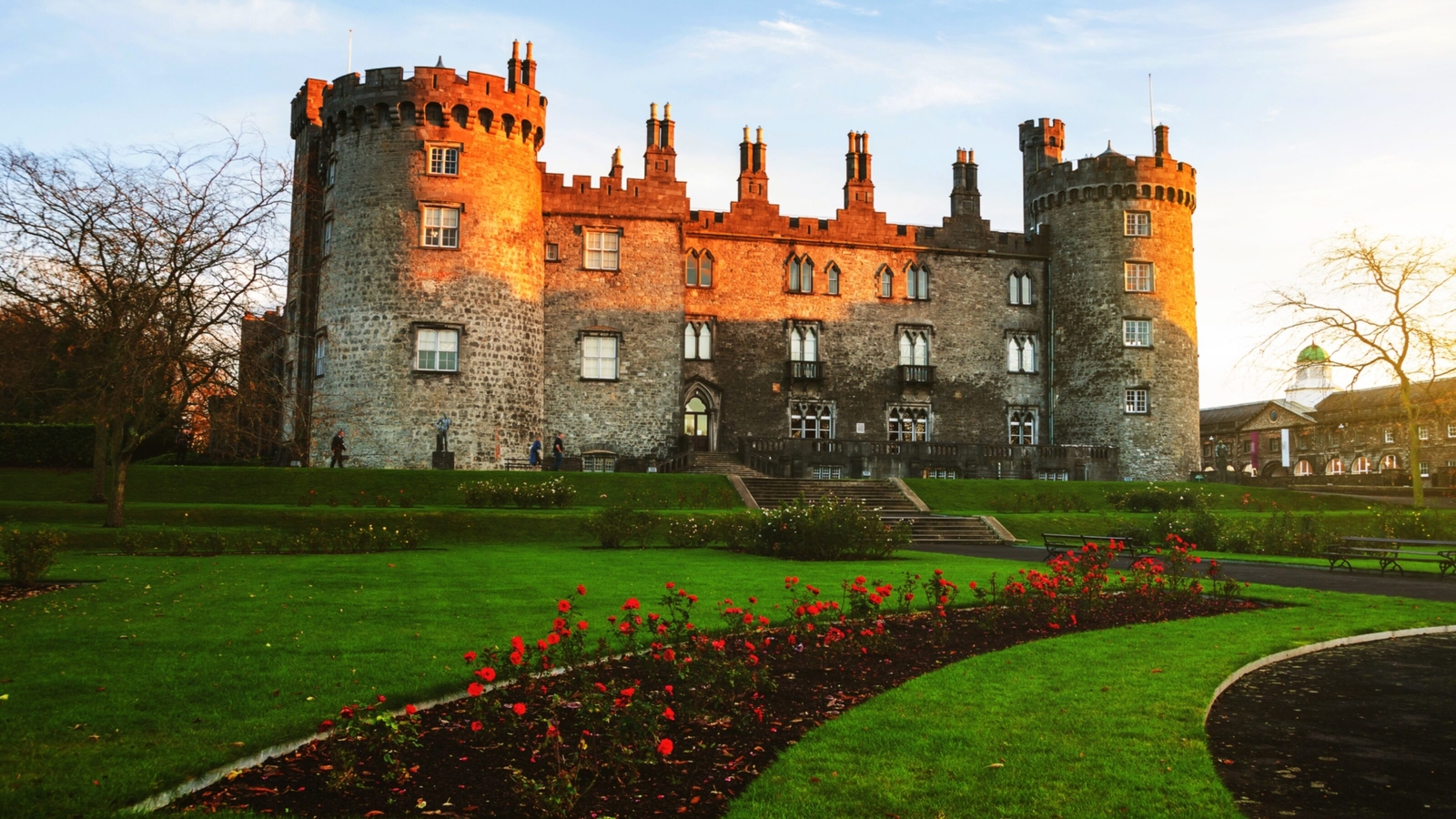 A historic stone castle with turrets bathed in golden sunlight, surrounded by vibrant green lawns and red flowers.