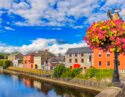 Colourful flowers atop a lamp post with a scenic view of a river and vibrant buildings under a blue sky.