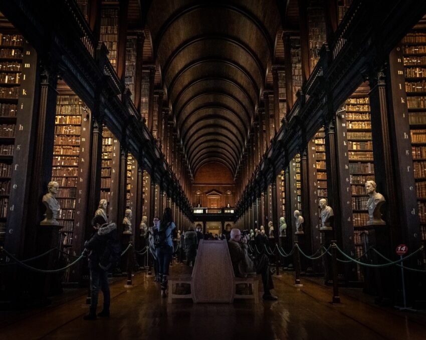 A grand library with towering bookshelves, statues, and visitors.
