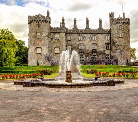 An ancient stone castle with towers behind a fountain and flowering garden.
