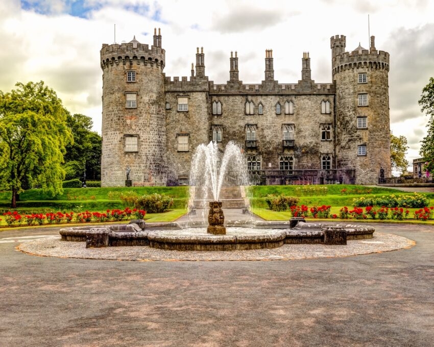 An ancient stone castle with towers behind a fountain and flowering garden.