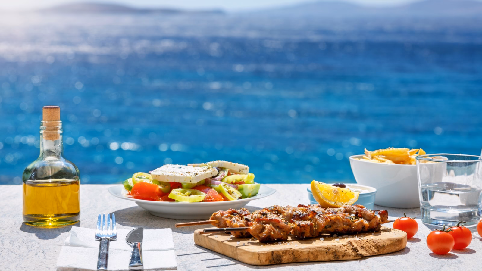 Greek meal with souvlaki, salad, fries, and olive oil, overlooking the sea.