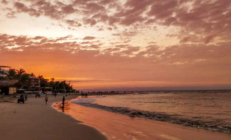 Sunset at the beach with people, reflecting water, and clouds in the sky.