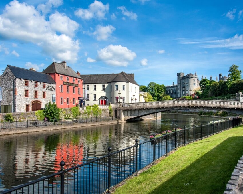 A scenic view of a river with an arched bridge, colourful buildings, and a castle in the background.