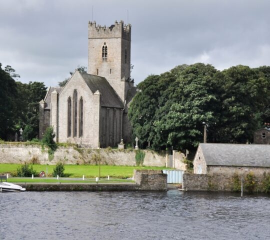 Historic stone church by a river with a boat and trees.