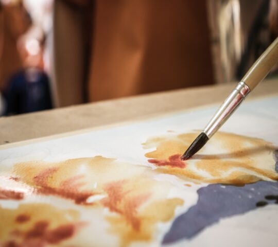 Close-up of a paintbrush applying watercolor on paper with blurred background.