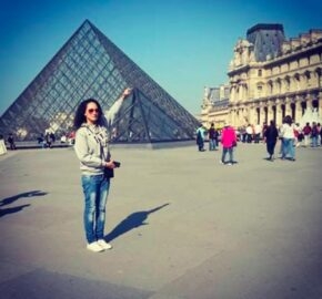 Person in front of the Louvre Pyramid, pretending to touch the tip, with other visitors around.