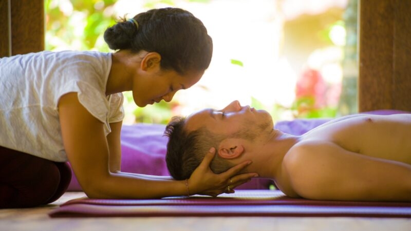 A person lying on a table indoors, appearing relaxed. The person is in comfortable clothing, and the setting suggests a peaceful atmosphere, possibly related to yoga or wellness activities.