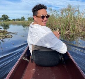 Person in white shirt canoeing in a river surrounded by reeds under a blue sky.