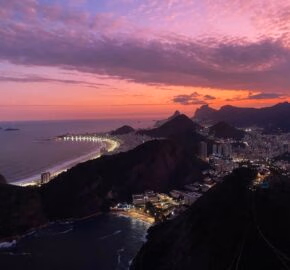 Aerial view of a coastal city at dusk with lit streets and a pink-hued sky.
