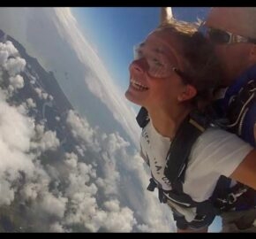 Two skydivers in tandem jump above clouds with mountains below.