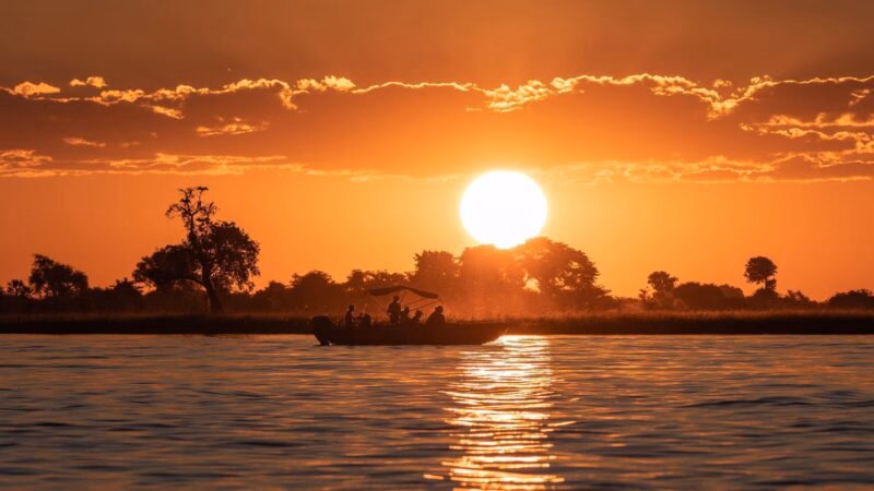 Sunset silhouette of a boat with people on a calm lake, with a large sun and orange sky in the background.