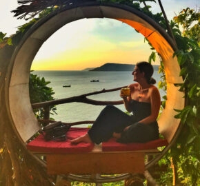 Person relaxing in a round wooden seat overlooking the sea at sunset.