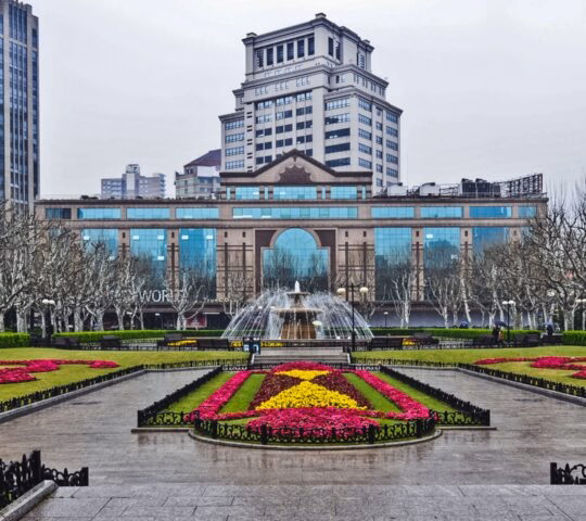 Urban park with colorful flower beds, a fountain, leafless trees, and a classical building background.