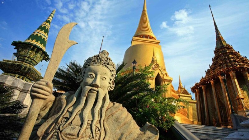 A traditional Thai temple with golden spires and lush greenery under a blue sky.