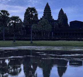 Angkor Wat silhouetted against a twilight sky, reflected in still water, with palm trees framing the ancient temple structure.