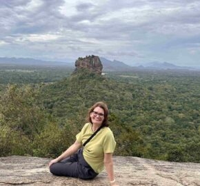 A person sits on a rocky ledge overlooking a lush green landscape with distant hills and a prominent rock formation under a cloudy sky.