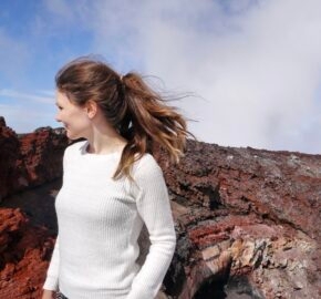 A woman in a white sweater stands near a volcanic landscape, with vibrant red and gray rock formations under a blue sky.