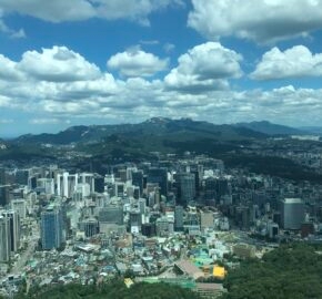 A panoramic view of a bustling cityscape with high-rise buildings, lush greenery, and distant mountains under a partly cloudy sky.