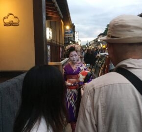 A traditional Japanese street scene at dusk, featuring a person in a colorful kimono walking past bystanders in a bustling area.