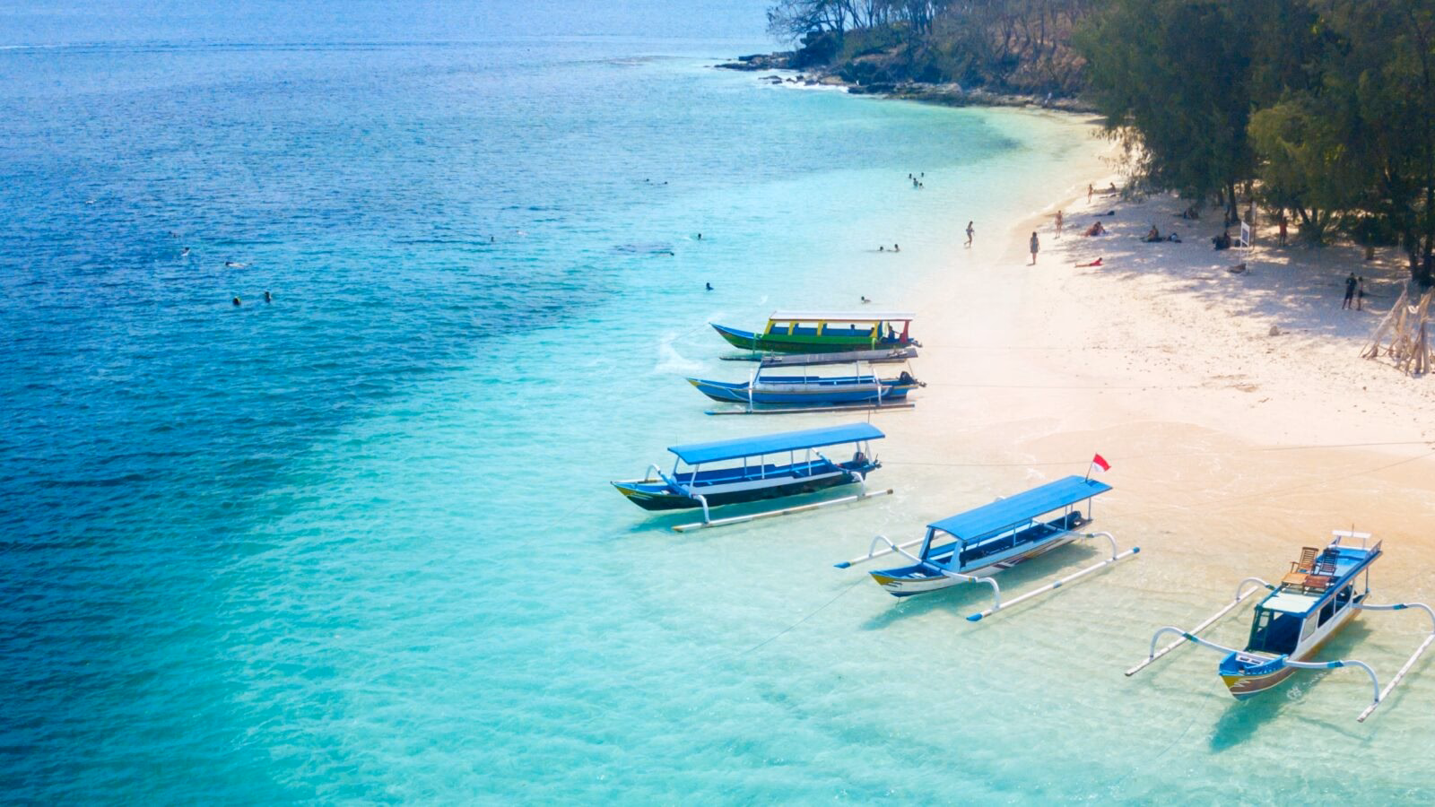Colorful boats anchored on a tranquil beach with clear turquoise water and people enjoying the sun and water activities.