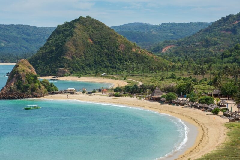 Scenic beach view with turquoise water, sandy shores, lush greenery, and hills in the background, dotted with small huts.