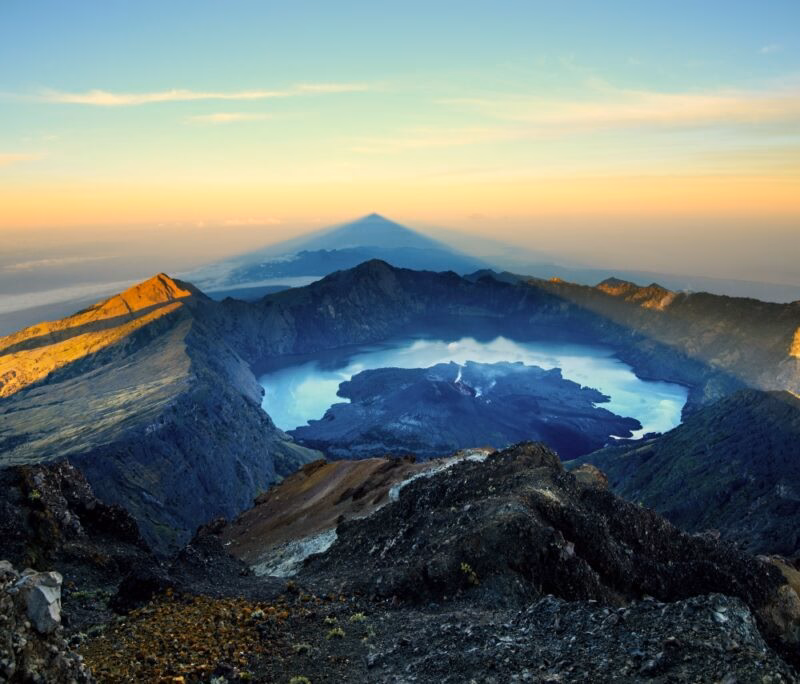 A stunning view of a volcanic crater at sunrise, featuring a tranquil blue lake surrounded by rugged mountains and soft shadows.