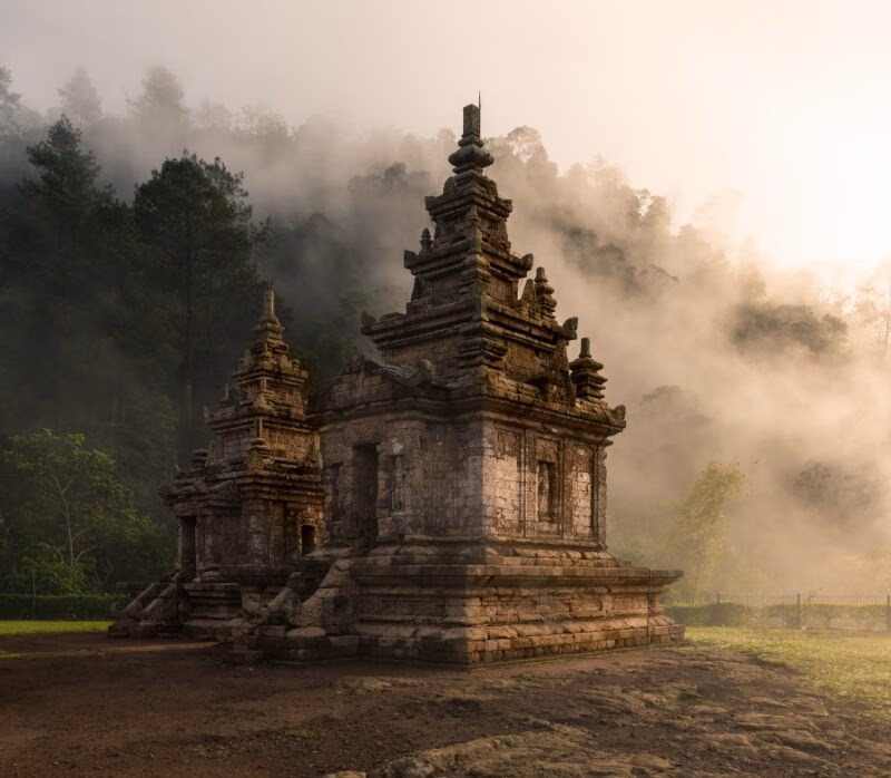 Ancient stone temple shrouded in mist, surrounded by lush greenery and silhouetted against a hazy morning sky.