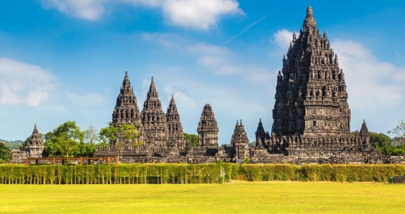 The majestic Prambanan Temple complex with multiple towering black stone structures against a blue sky and green landscape.