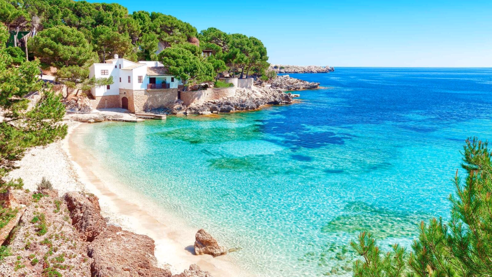 A tranquil coastal scene featuring a white house nestled among lush greenery, with crystal-clear turquoise waters and a sandy beach.