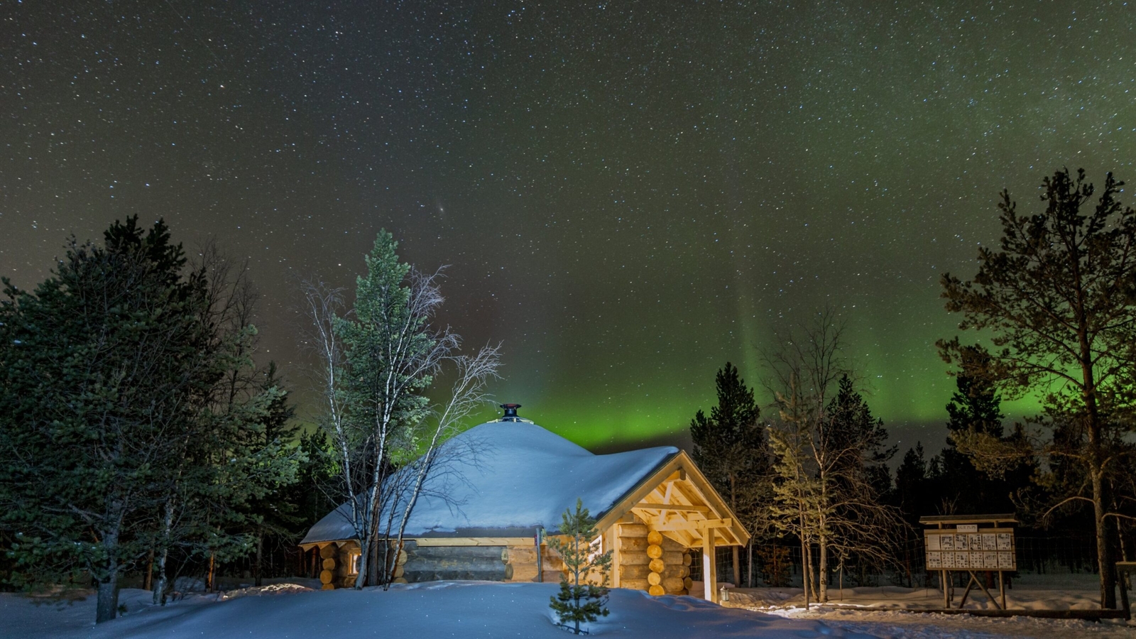 A cozy log cabin covered in snow, surrounded by trees under a starry night sky with green auroras illuminating the horizon.