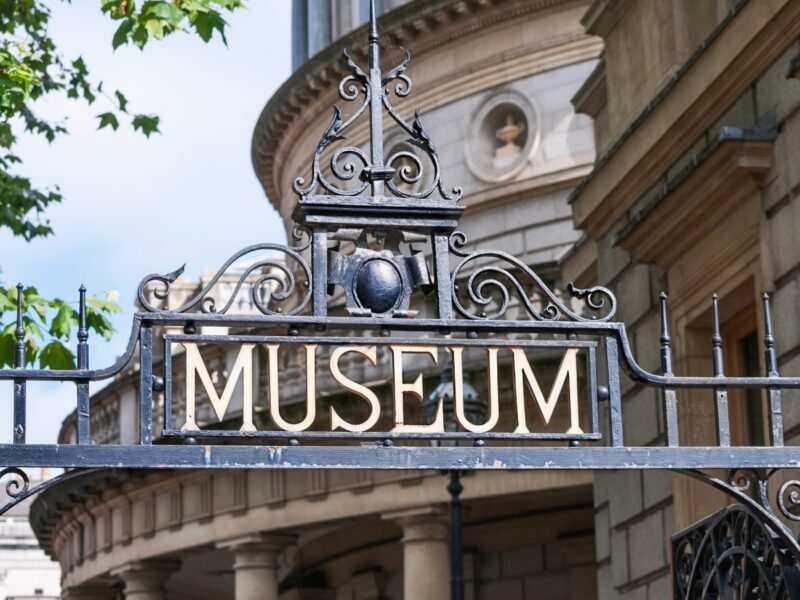 Close-up of an ornate black iron gate with the word "MUSEUM" prominently displayed, framed by green leaves and classical architecture.