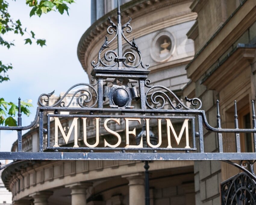 Close-up of an ornate black iron gate with the word "MUSEUM" prominently displayed, framed by green leaves and classical architecture.