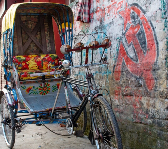 A colourful rickshaw parked against a graffiti-covered wall, featuring vibrant seating and a vintage design.
