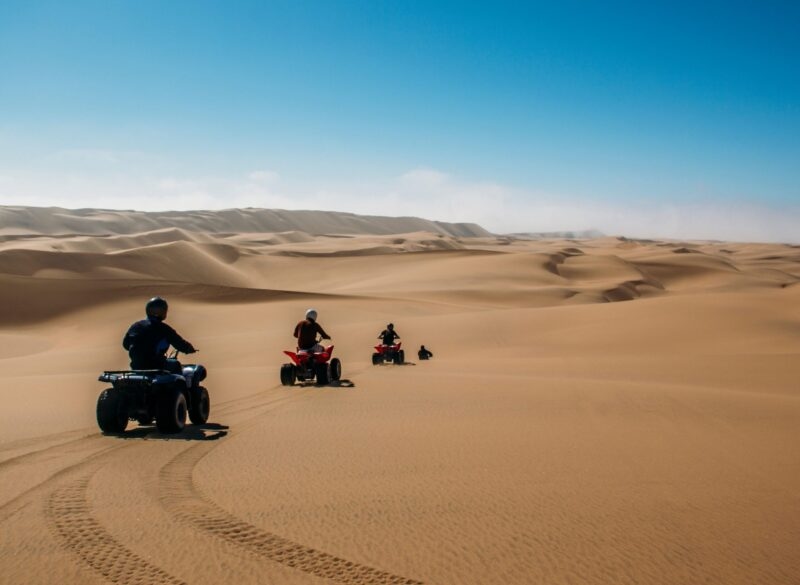 Four people riding quad bikes across expansive sand dunes under a clear blue sky during Namibia family tours.
